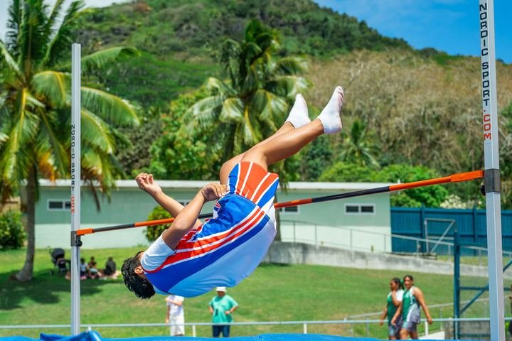 Clearing the bar in the high jump, with the hills of Rarotonga behind.